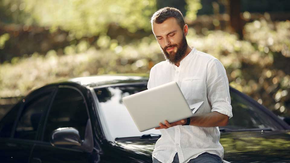 Ein Mann steht neben seinem Auto und nutzt einen Laptop, um das Auto online zu verkaufen